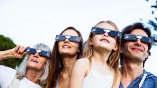 a family look up toward the sun wearing eclipse glasses, they are all smiling.