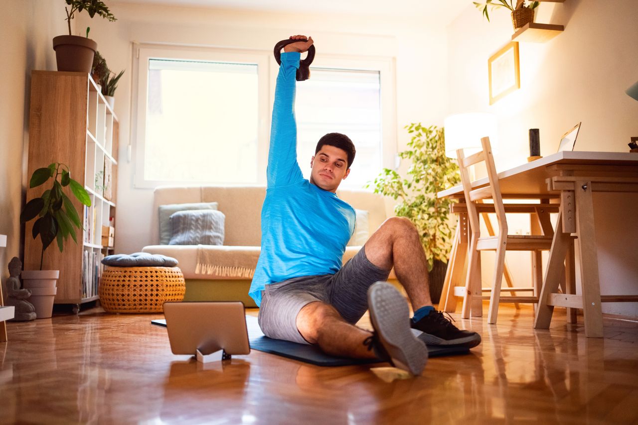 Young man exercising turkish get-up with a kettlebell, online workout on tablet in his living room