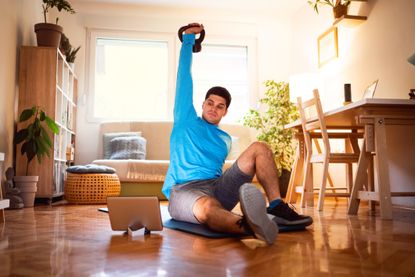 Young man exercising turkish get-up with a kettlebell, online workout on tablet in his living room