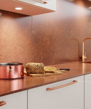 A kitchen with a copper splashback and countertop, a copper saucepan, a plate of bread, and white cabinets with copper handles