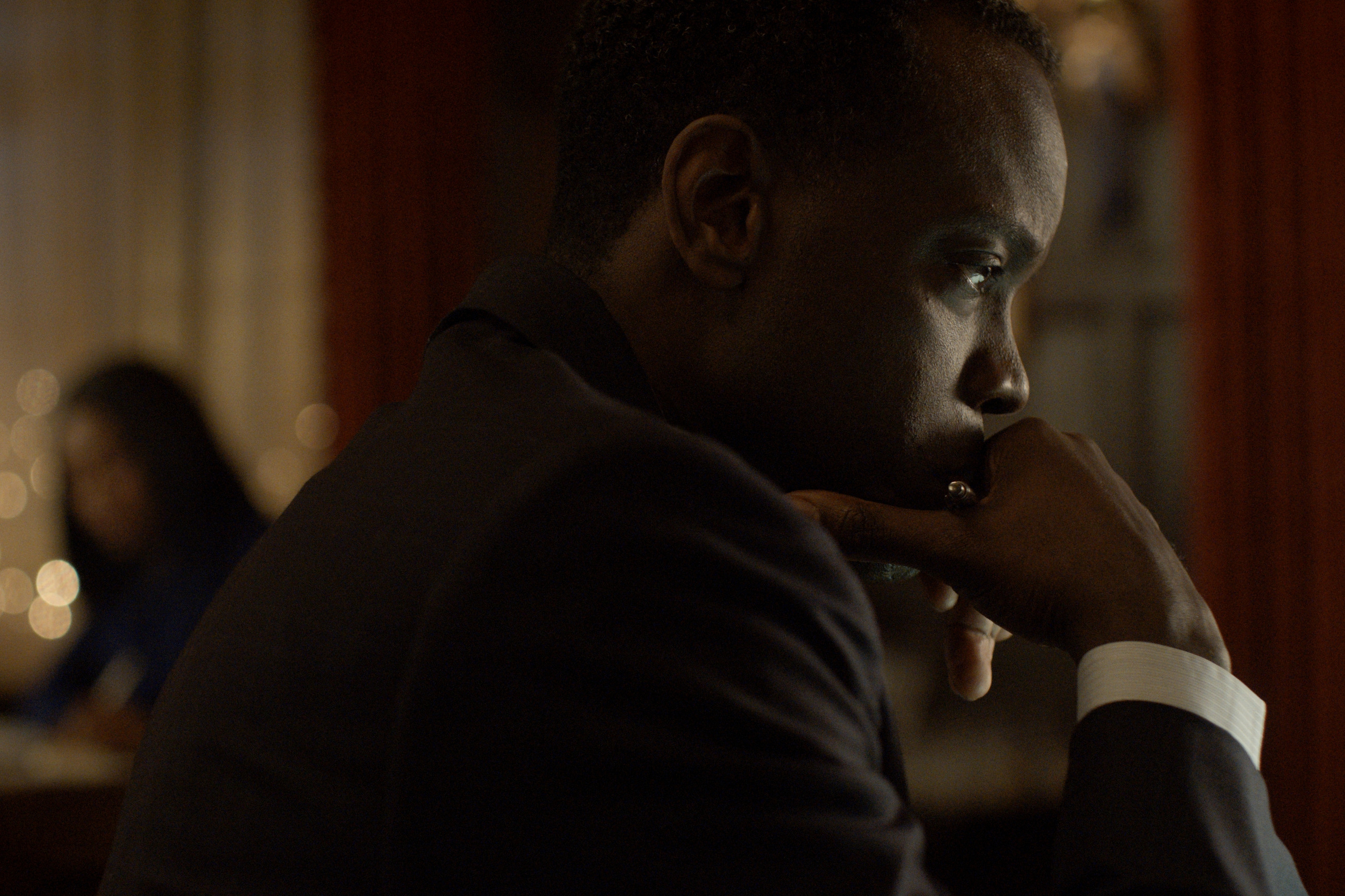 David Gyasi wearing a suit and resting his chin in his hand in a still from the diplomat season 3