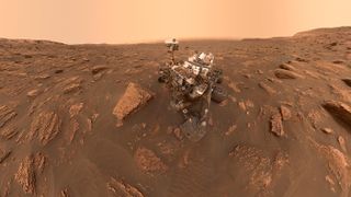 curiosity mars rover on the surface of mars, surrounded by boulders. behind is a peach, dusty sky