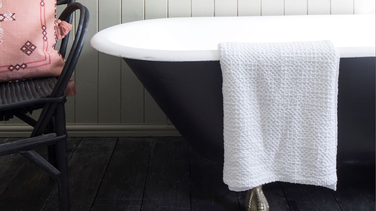 Black bathtub with towel hanging over, black rattan chair with cushion