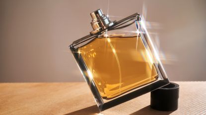 Image of a sunlit perfume bottle filled with gold liquid, stood at an angle on the lid and set against a wood and taupe wall background