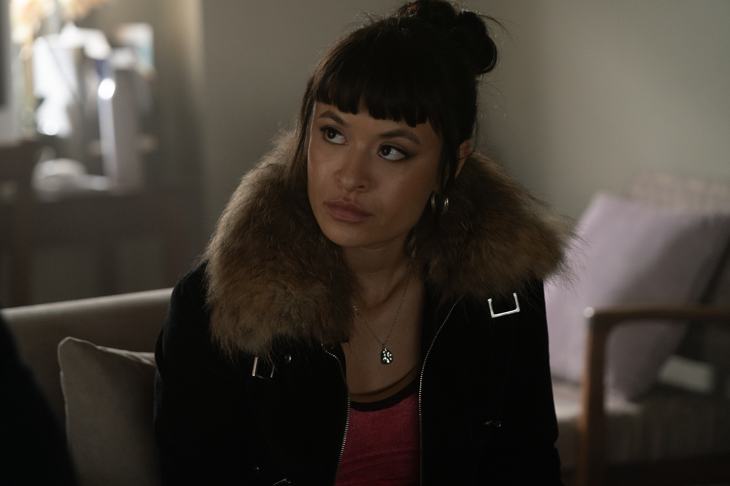 sonia mena as pippa wearing a fur lined coat sitting in a waiting room with her hair in a bun in a still from tell me lies season 3