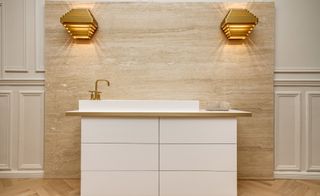A sink with brass tap in front of an ivory-coloured wall with two lights in brass fixtures