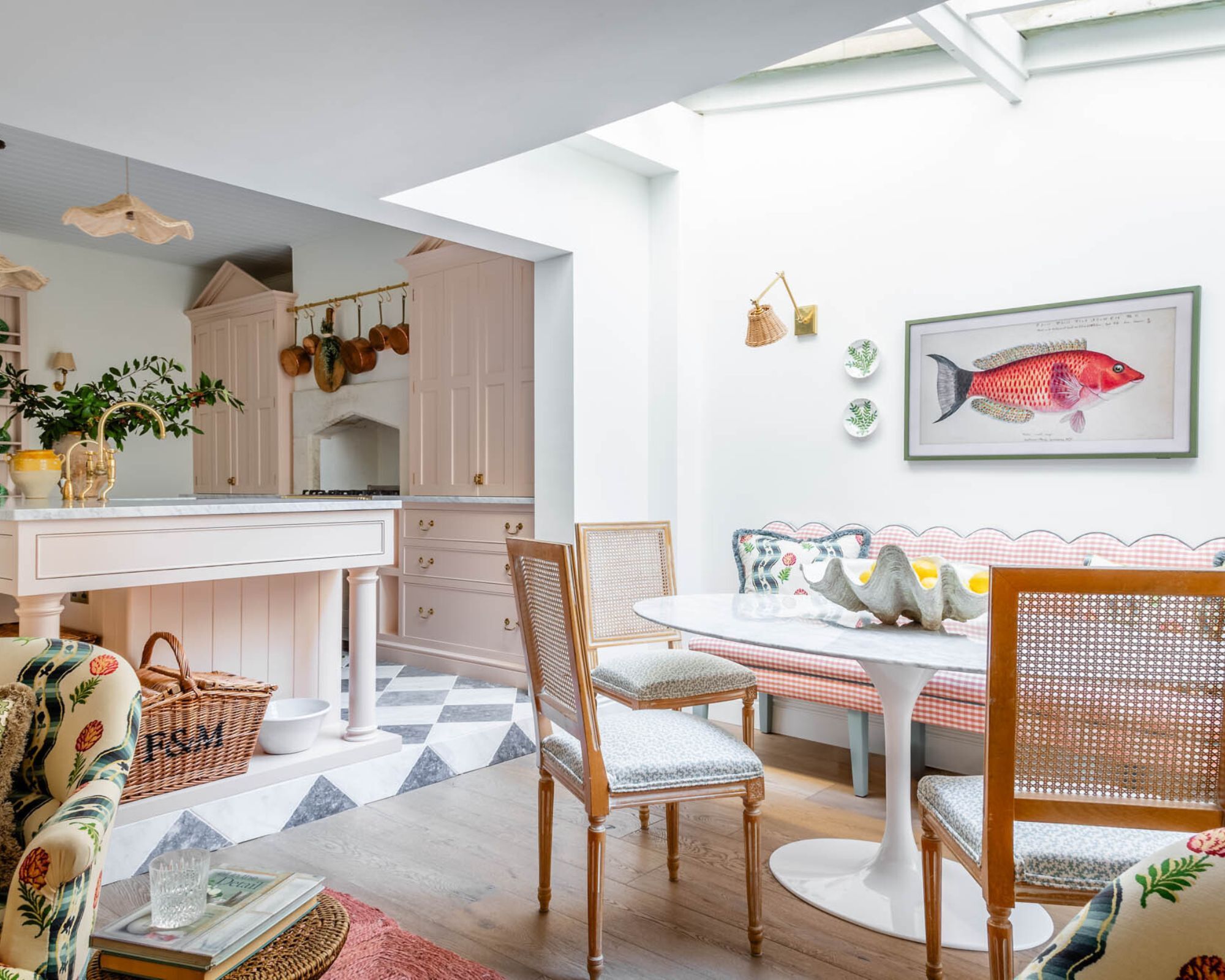 A large pale pink kitchen with a pink kitchen island, next to this is a dining area with a white table and three rattan chairs. A bright and colourful painting on the wall of a pink fish.