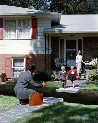 Stereotypical midcentury scene of man arriving home from work, being greeted by wife and children. From the exhibition Suburbia. Building the American Dream