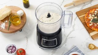 The Oster 10-Cup Food Processor with Easy-Touch Technology making pizza dough from scratch next to ingredients