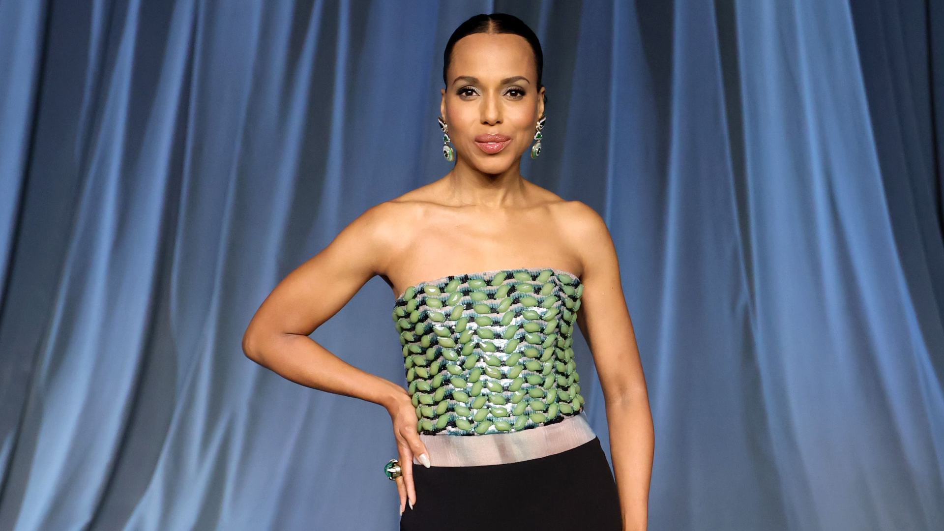 Kerry Washington wearing a slicked bun hairstyle and strapless gown. She has her hand on her hip and is smiling at the camera.
