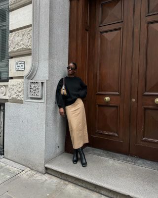 Woman wears black sweater, beige skirt and black boots