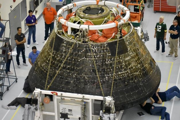 A large black-charred space capsule sits in a white room, elevated on a large white hoister. two individuals on the right are laying underneath, their black shirts and blue jeans sticking out. Other people stand around loitering.