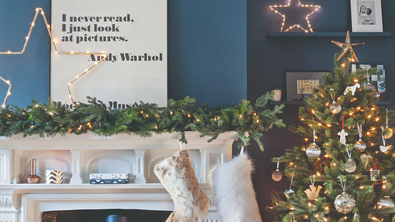 A blue-painted living room with a white fireplace decorated for Christmas with festive lights and a Christmas tree next to it