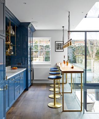 Mezzanine kitchen with blue cabinets and dark wooden floor, kitchen bar with metallic frame and exposed wooden countertop with jagged edge, rounded bar stools with metal frames and blue upholstered seat