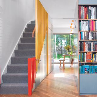 hallway with storage and bookcase and doors painted in duck egg blue, with staircase opposite with grey stair carpet and yellow and orange painted bannisters