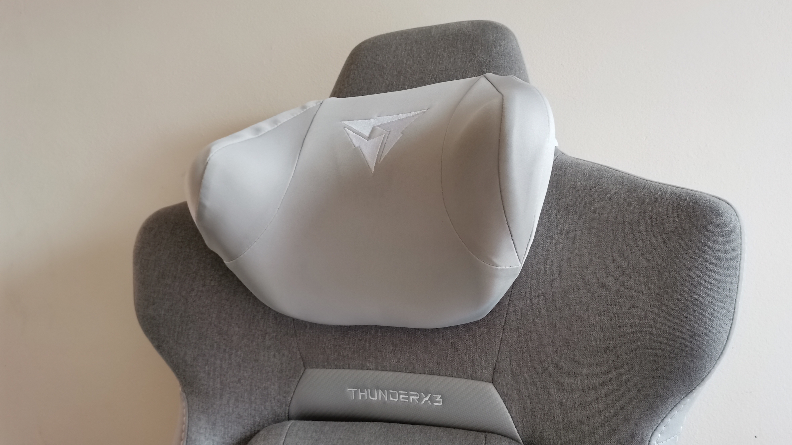 The ThundercoreX3 Core gaming chair in grey in front of a pale wall, next to a wooden bed frame