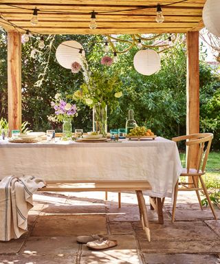 Dining table covered in linen tablecloth sat outside on patio underneath wooden pergola