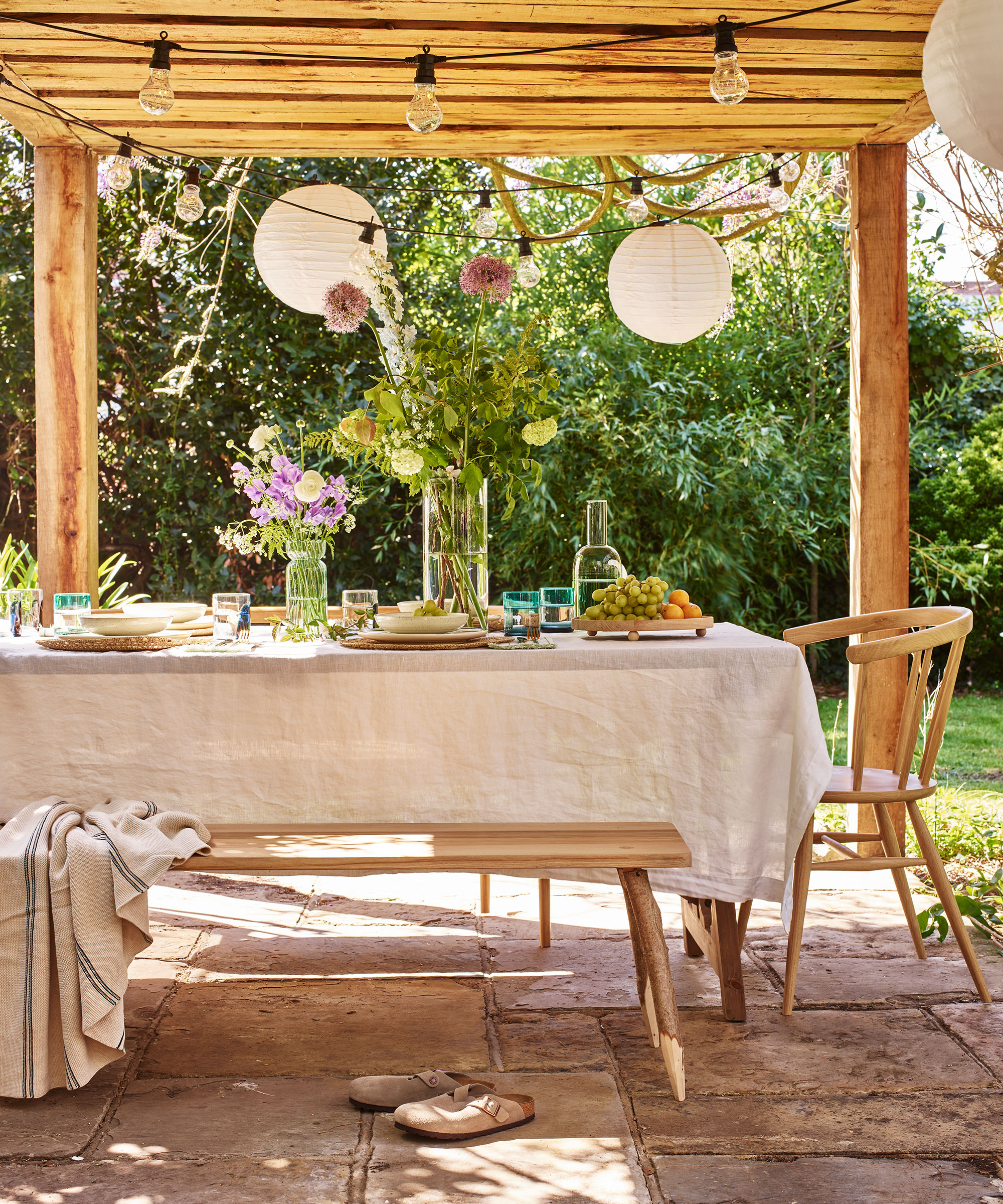 Dining table covered in linen tablecloth on patio underneath wooden pergola with string lights and paper lanterns