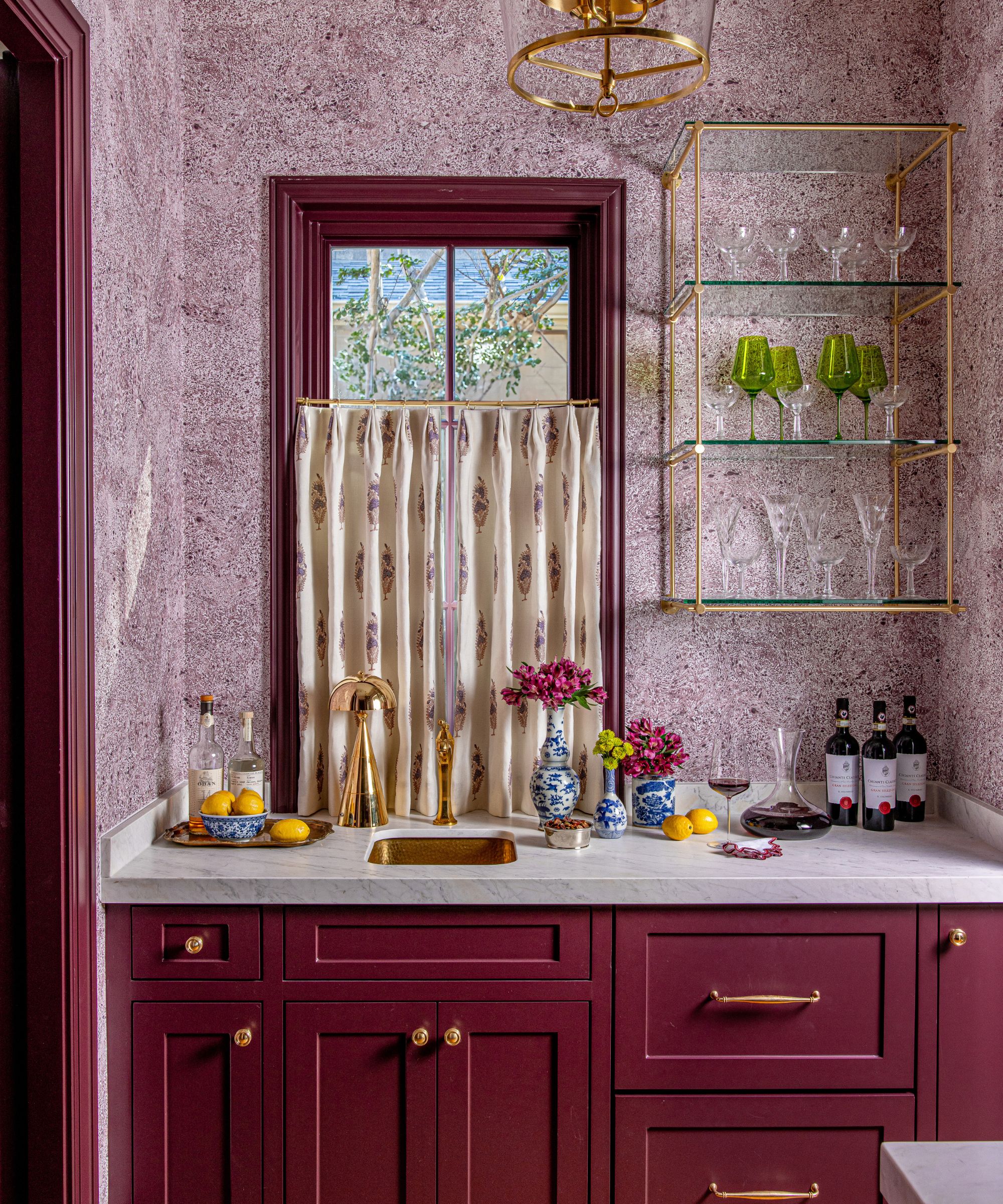 Home bar with burgundy painted cabinets and window frame, gold hardware, marble countertop and sink and open shelving with glasses