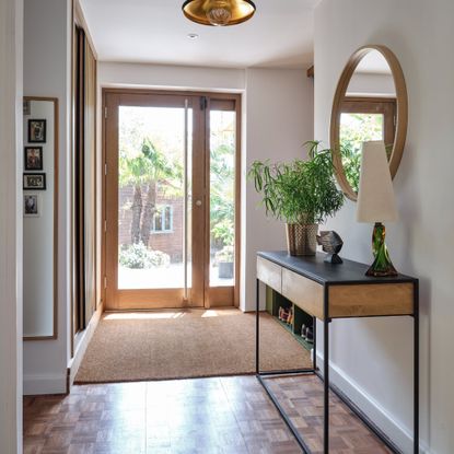 A hallway with a glass front door and a jute rung in front of a shoe storage bench and a console table with a round wall mirror hung above it
