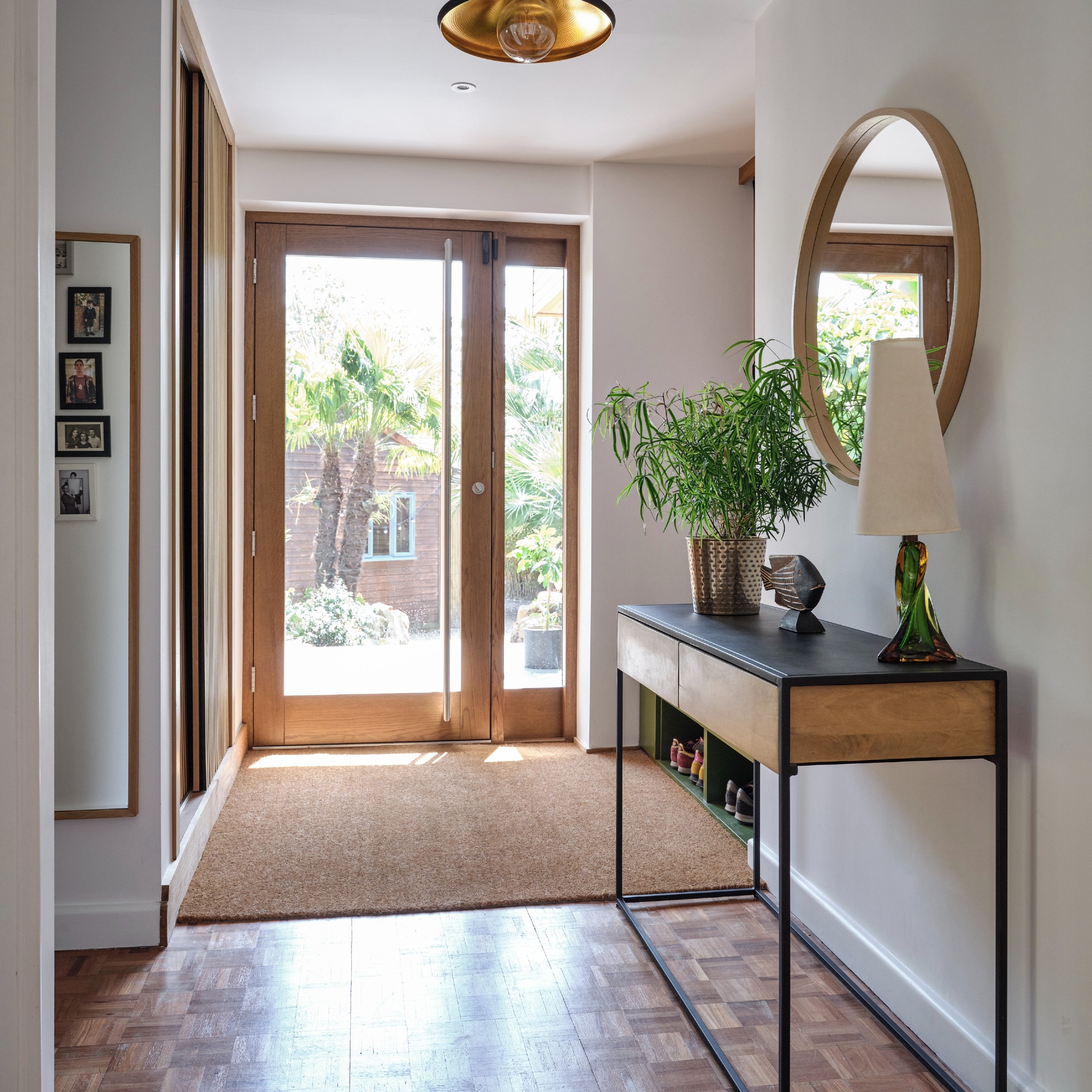 A hallway with a glass front door and a jute rung in front of a shoe storage bench and a console table with a round wall mirror hung above it