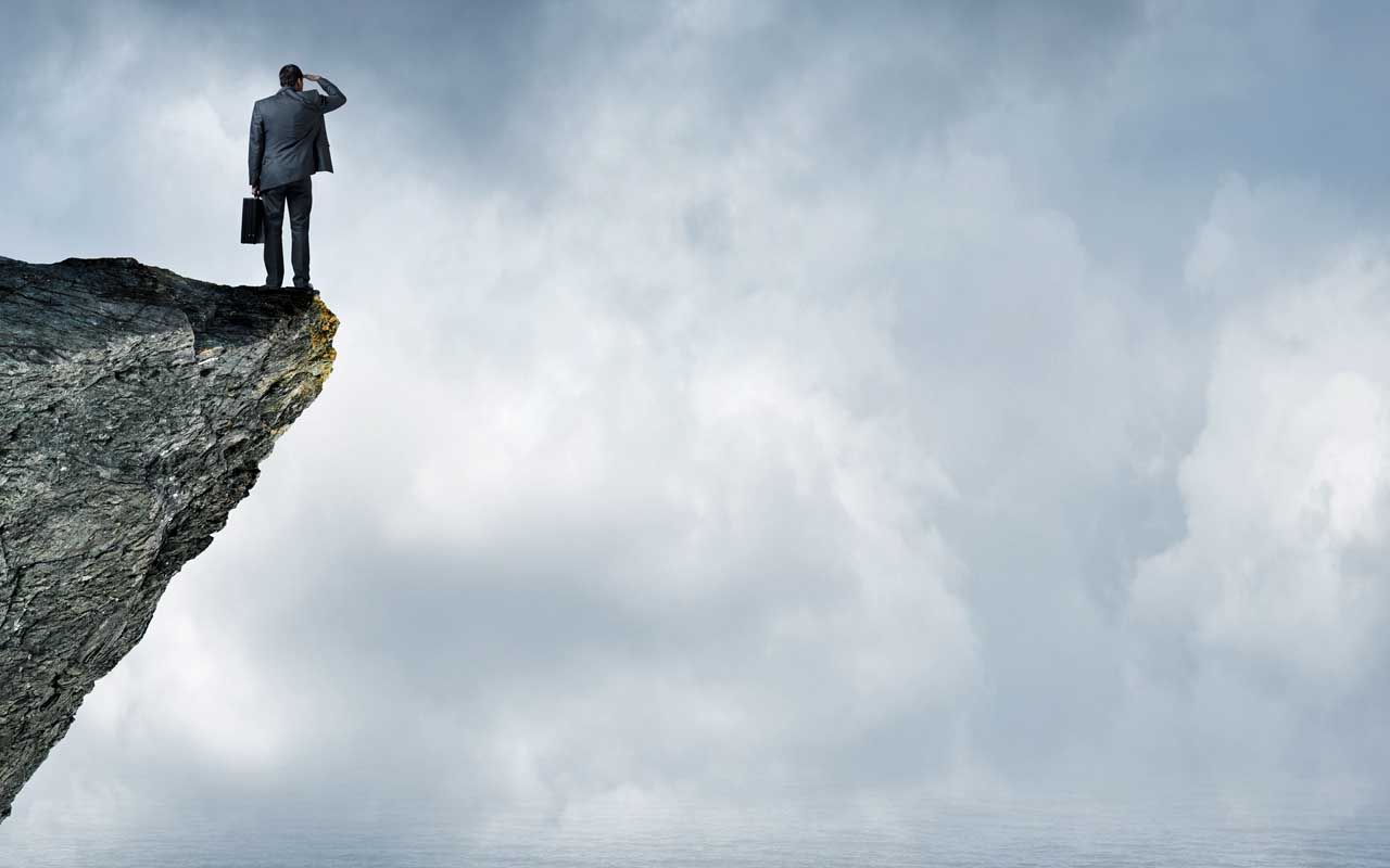 A businessman standing on the edge of a cliff and holding his briefcase, peers out towards a fog shrouded sea.Ample copy space is created by the clouds.