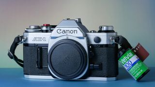 Cámara Canon AE-1 sobre una superficie azul y fondo pastel junto a un rollo de película Fujifilm Velvia