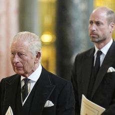 Prince William and King Charles wearing black suits looking somber