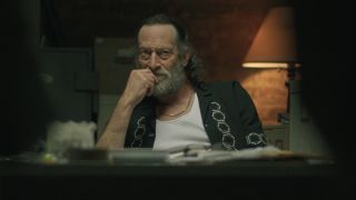 Troy Kotsur in Black Rabbit putting his left hand up to his chin with his elbow resting on the table. 