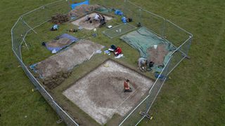 This photo shows an overview of an excavation site of a 10,000-year-old pit near Stonehenge. A large rectangle area is fenced off and inside there are 2 sites dug up. There are a team of 7 archaeologists working on this site.