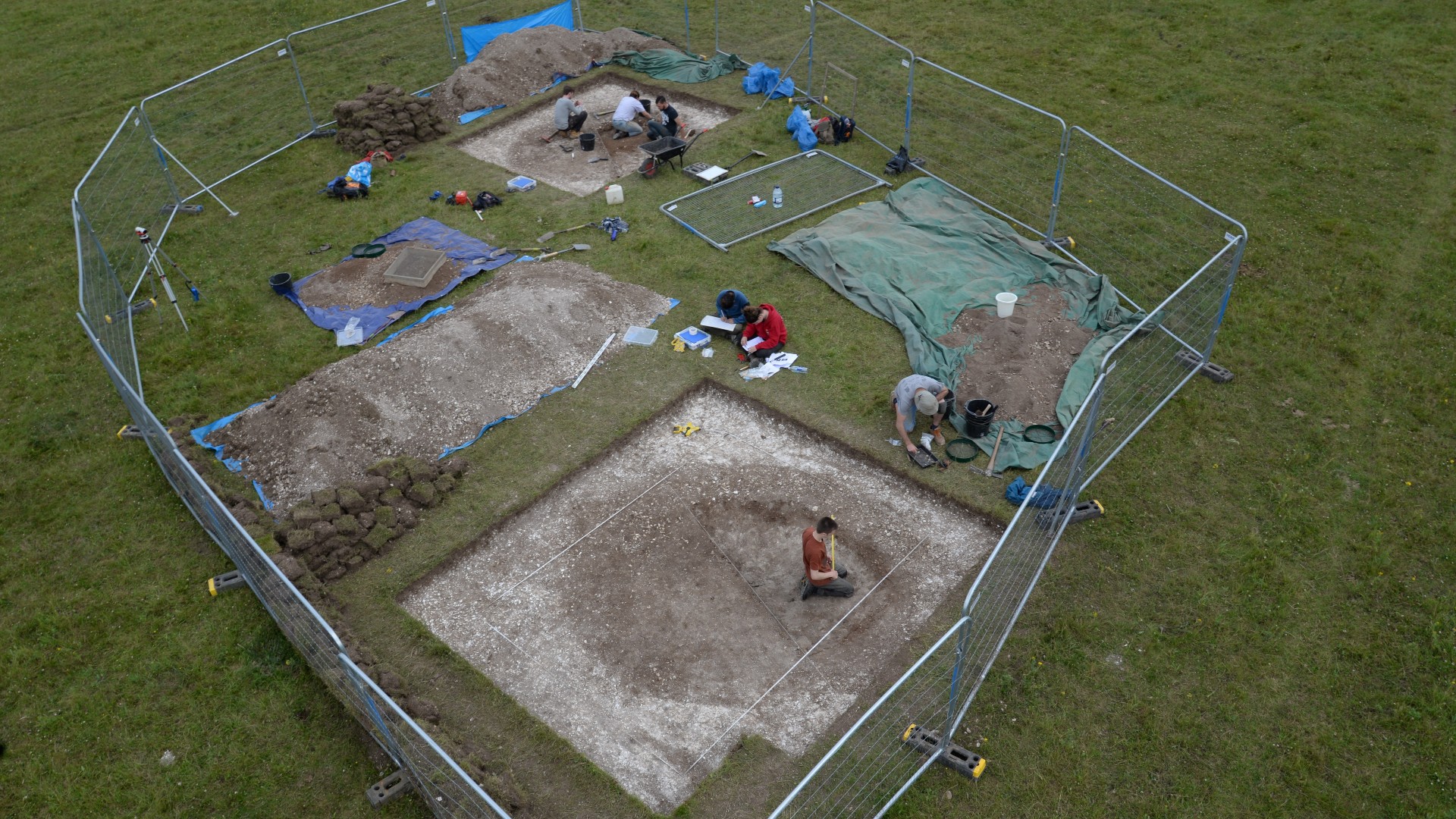 Deze foto toont een overzicht van een opgravingslocatie van een 10.000 jaar oude put in de buurt van Stonehenge.  Een groot rechthoekig gebied is omheind en binnen zijn er 2 locaties opgegraven.  Er is een team van 7 archeologen aan het werk op deze site.