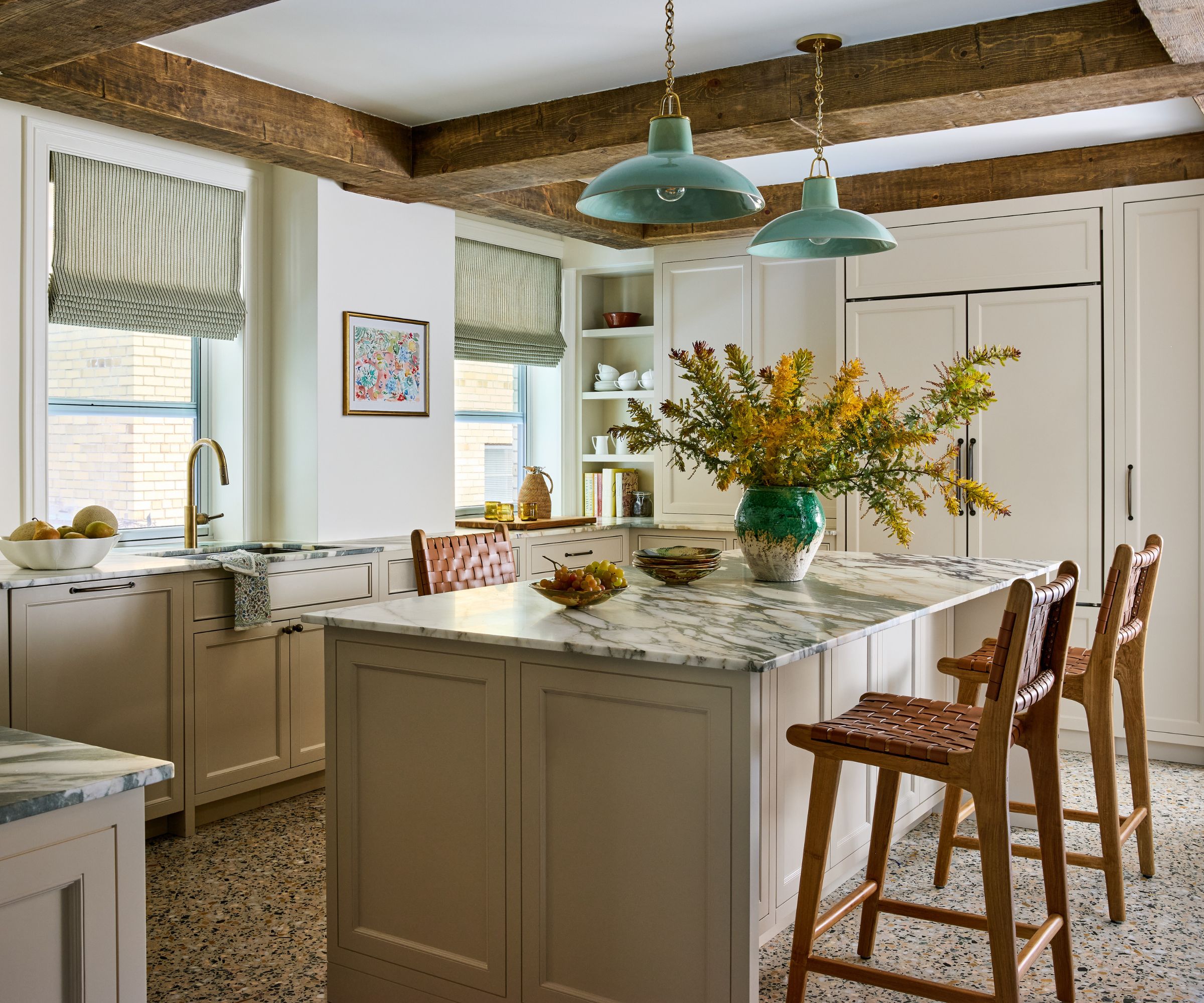 A kitchen with neutral cabinets, white walls, marble counters, and a kitchen island with two tan bar stools and wood ceiling beams.