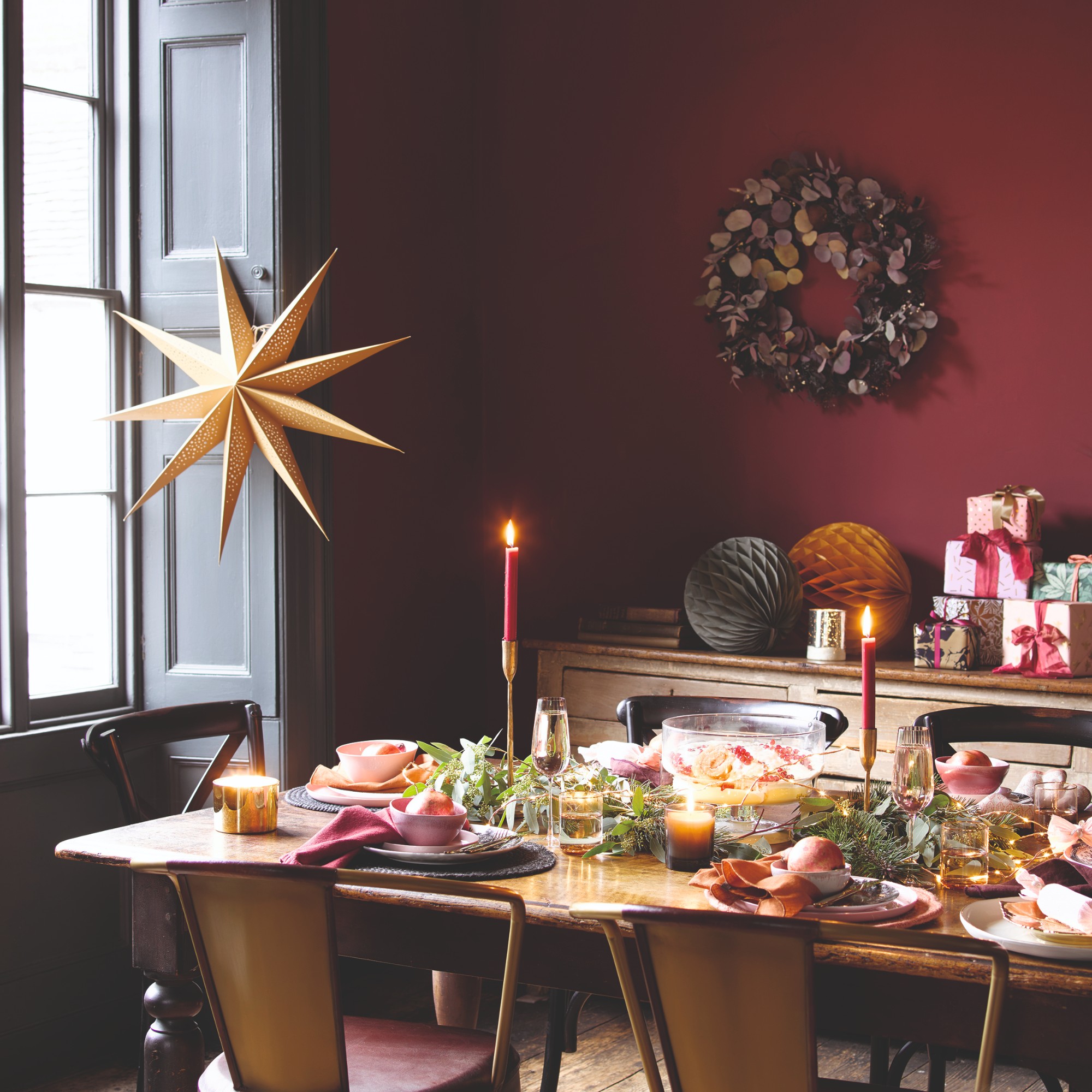 A burgundy-painted dining room decorated for Christmas with a wreath and a paper star on the window and a set table