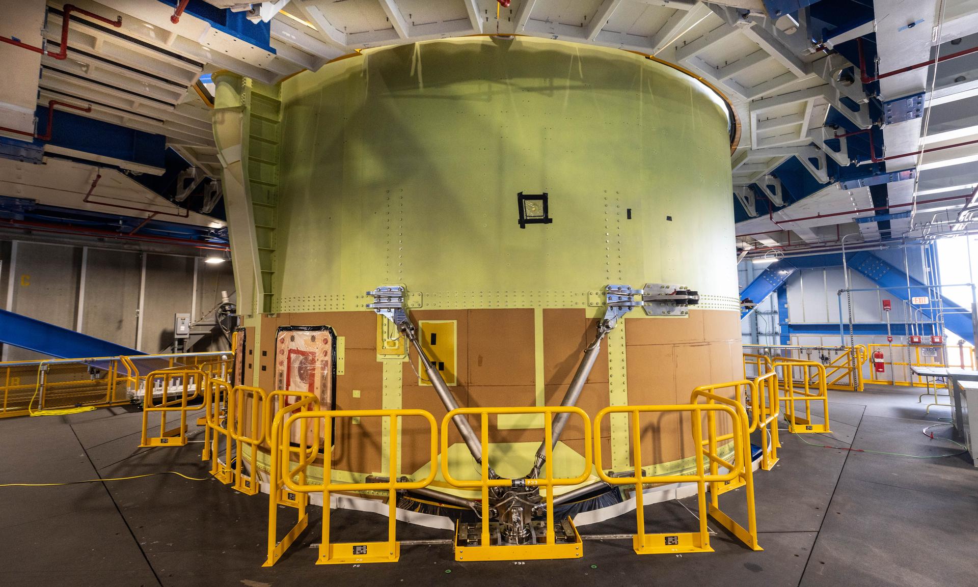 A large yellow cylinder stands from concrete floor to white metal ceiling, surrounded by temporary yellow railing in a warehouse setting. Teams from Kennedy lift NASA&amp;rsquo;s integrated Artemis 3 SLS core stage engine section with its boat-tail inside the center&amp;rsquo;s Vehicle Assembly Building at NASA&amp;rsquo;s Kennedy Space Center in Florida on Aug. 13, 2025. Shown inside the facility&amp;rsquo;s High Bay 2 for processing,