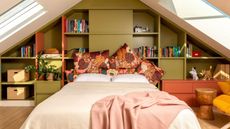 attic bedroom with green cabinetry on wall behind a bed with colorful cushioned bedhead, white pillowcases, cream sheets with a pink throw draped across the bottom, with skylights