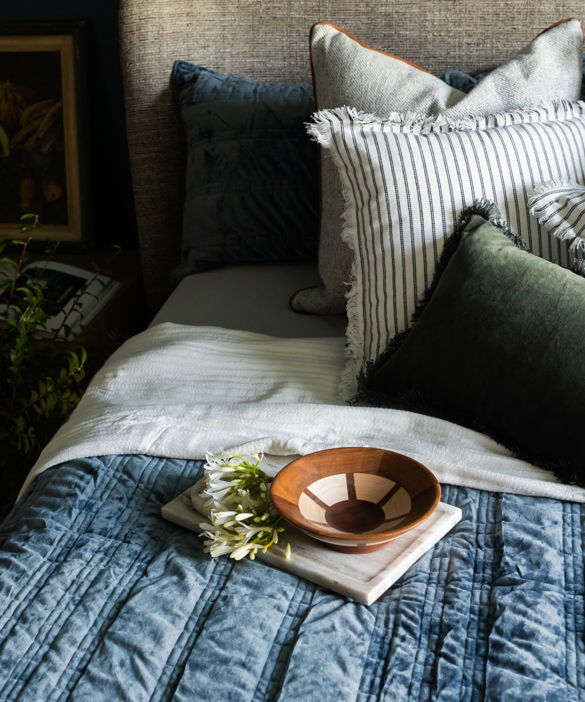 Closeup of made bed with a padded blue duvet and an assortment of cushions, with a small bowl and flowers positioned on the bed.