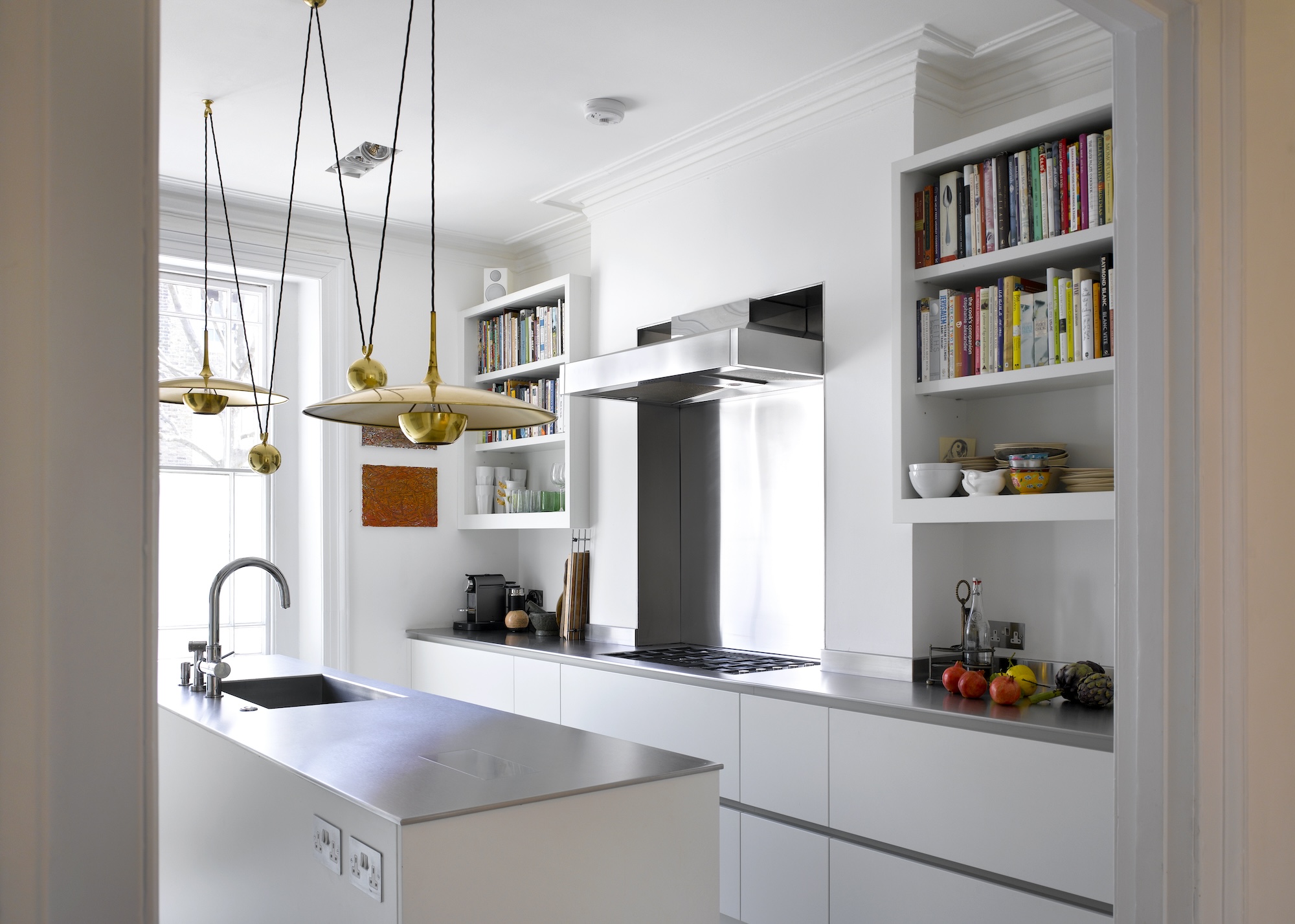 A stainless steel kitchen with large pendant lights hanging above. There is a view of the stove with a large extraction fan above it and on either side of the stove are shelves where books and kitchen essentials have been place