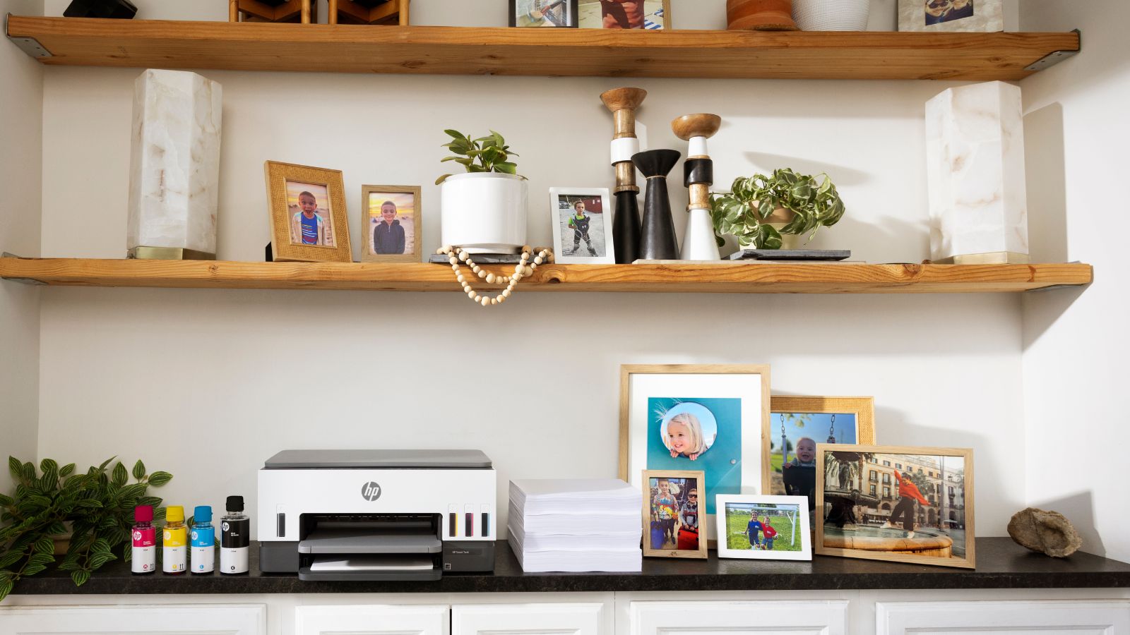 HP Smart Tank printer sits on a shelf with printed family photos and a stack of paper plus bottles of ink.
