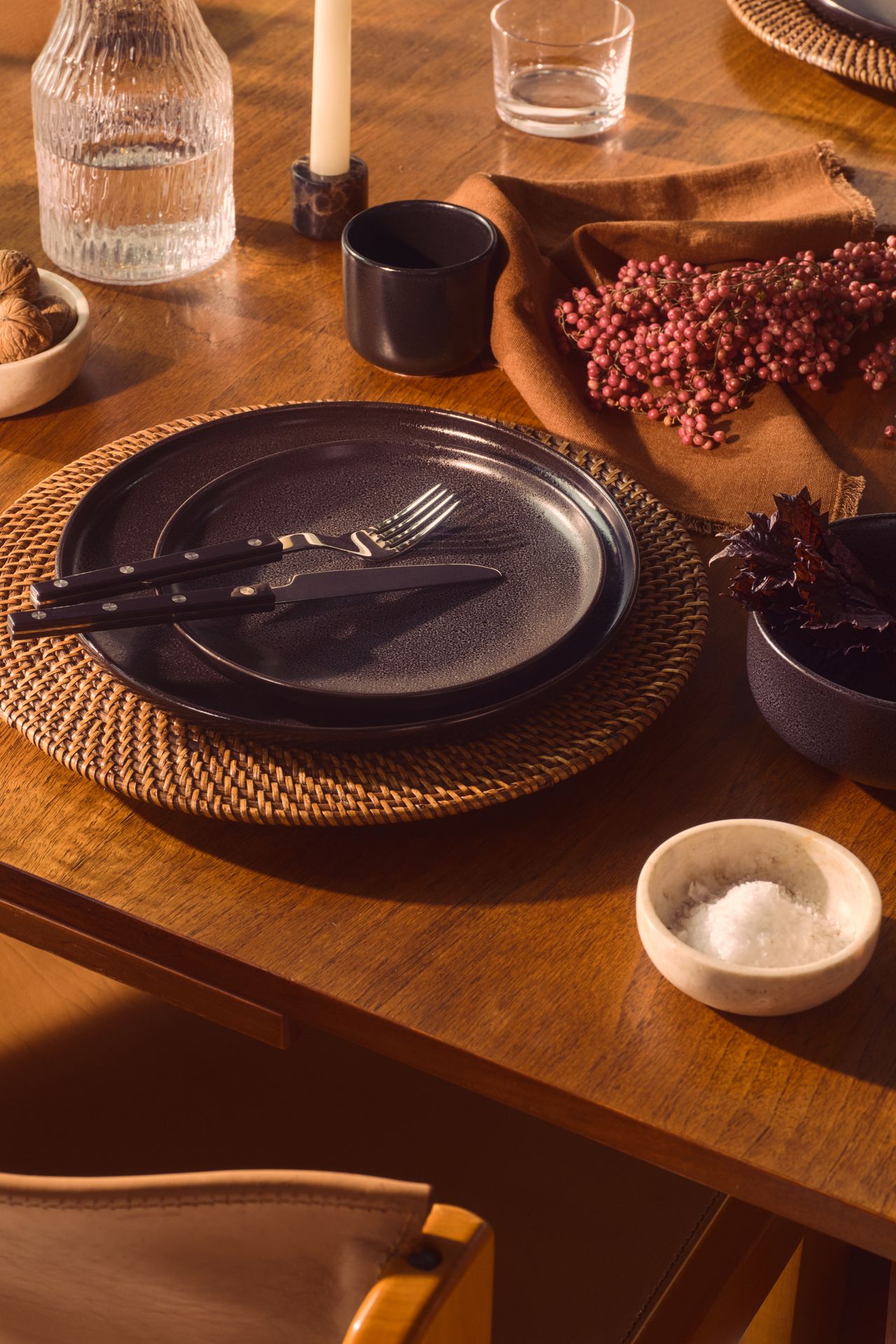 Close-up image of a place setting on a wooden dining table. There is a rattan charger plate with two black dinner plates stacked on top of it. There are orange napkins on the tables as well as a few glassware items and ceramic bowls.