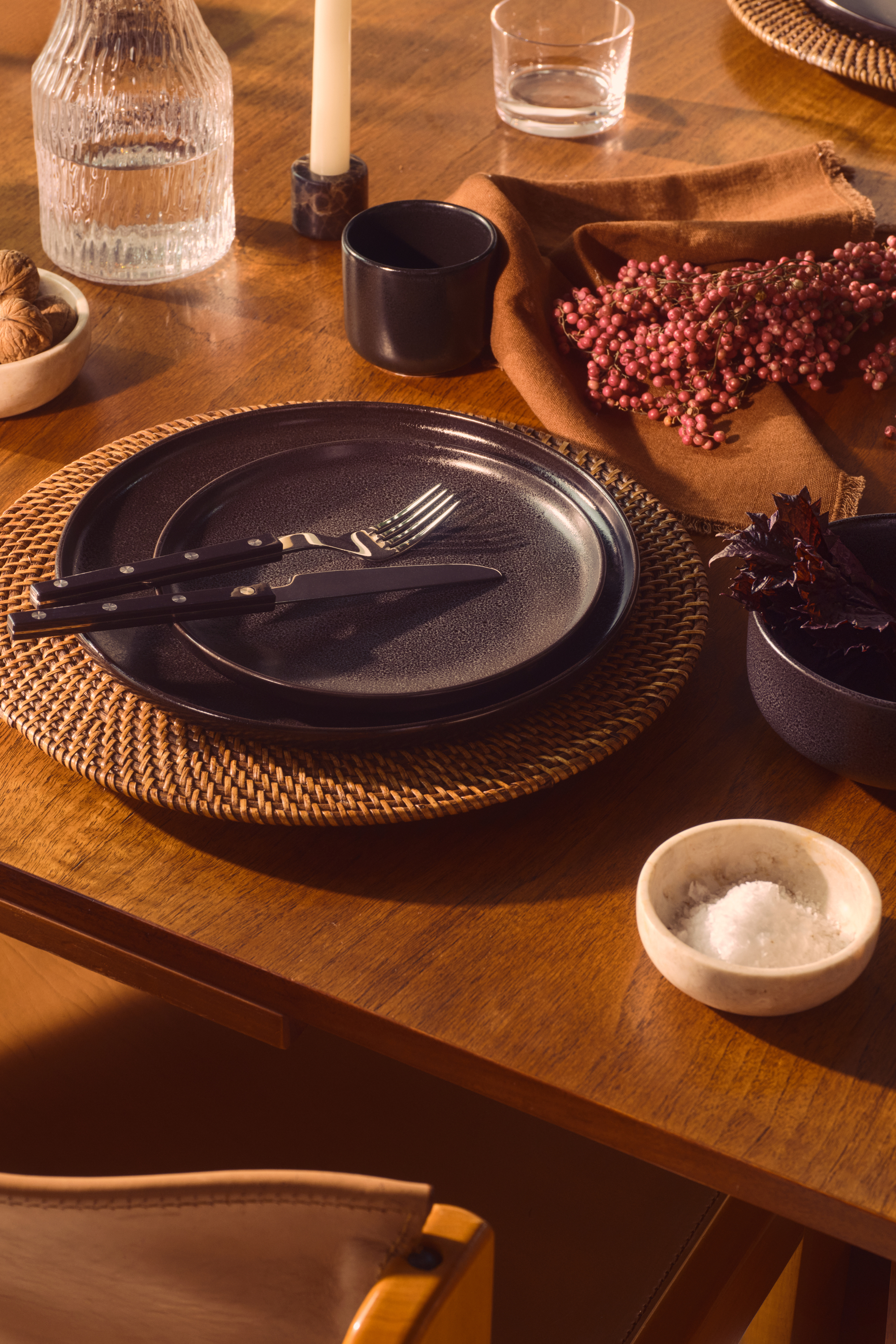 Close-up image of a place setting on a wooden dining table. There is a rattan charger plate with two black dinner plates stacked on top of it. There are orange napkins on the tables as well as a few glassware items and ceramic bowls.