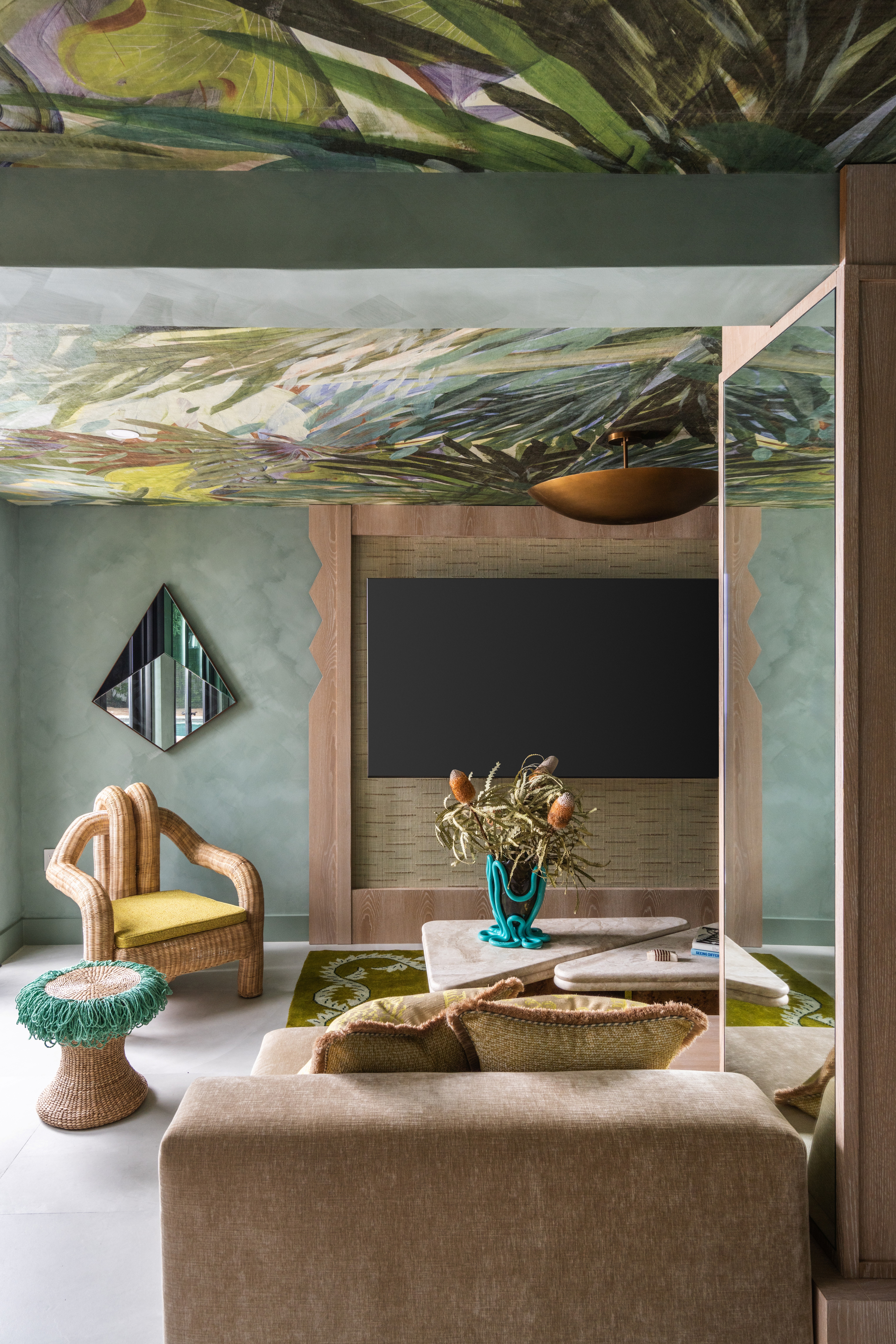Interior of a tropical home with teal walls, zellige tiles surfaces, wavy, sand-coated details, rattan furniture mixed with bright aquamarine and ochre touches, and a botanical-themed mural ceiling.