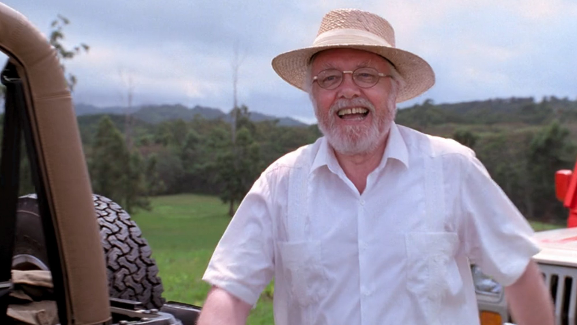 Sir Richard Attenborough as Hammond, smiling in a white shirt and brown hat in Jurassic Park, just as the people see the dinosaurs for the first time.