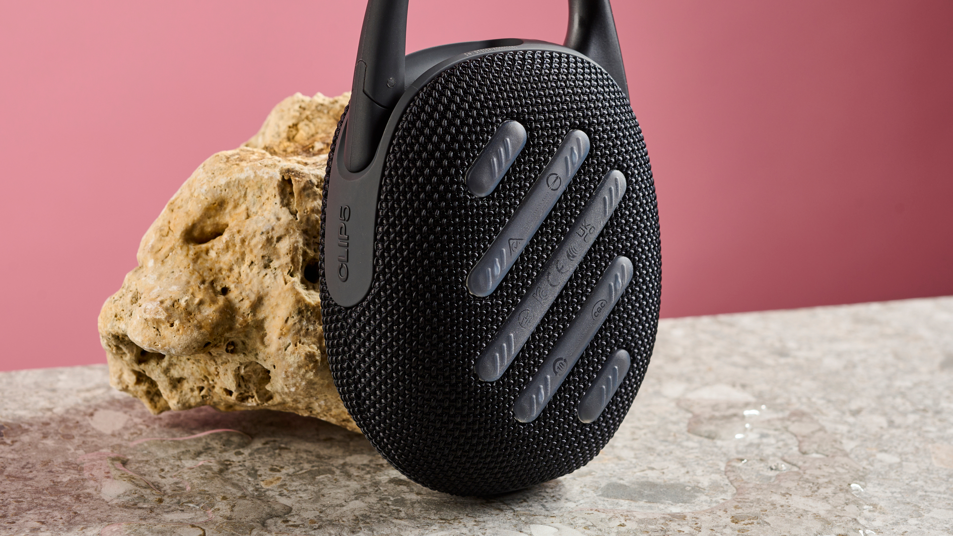 A close up of the back of the JBL Clip 5, pictured leaning against a sand coloured rock against a pink background,