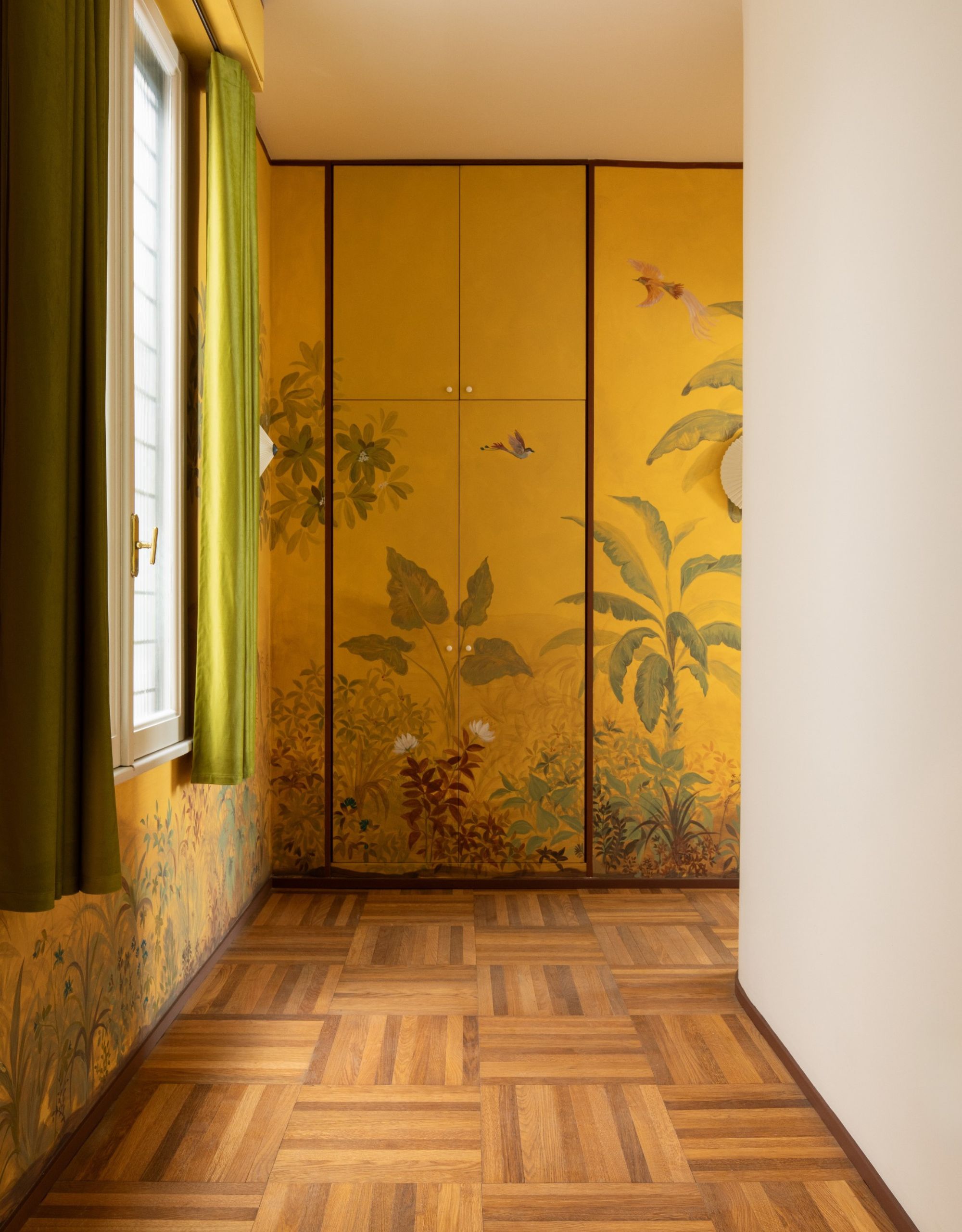 A yellow-painted hallway mural conceals cubpoards next to a window with green curtains alongside a white wall on the other side, a white ceiling, and wood flooring