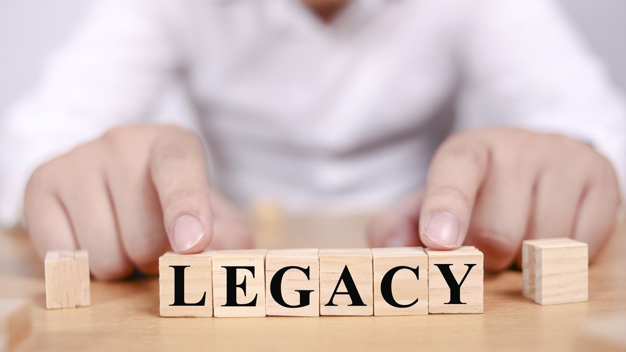 A man rests his index fingers on the L and the Y in the word &quot;legacy,&quot; which is spelled out in wooden blocks.