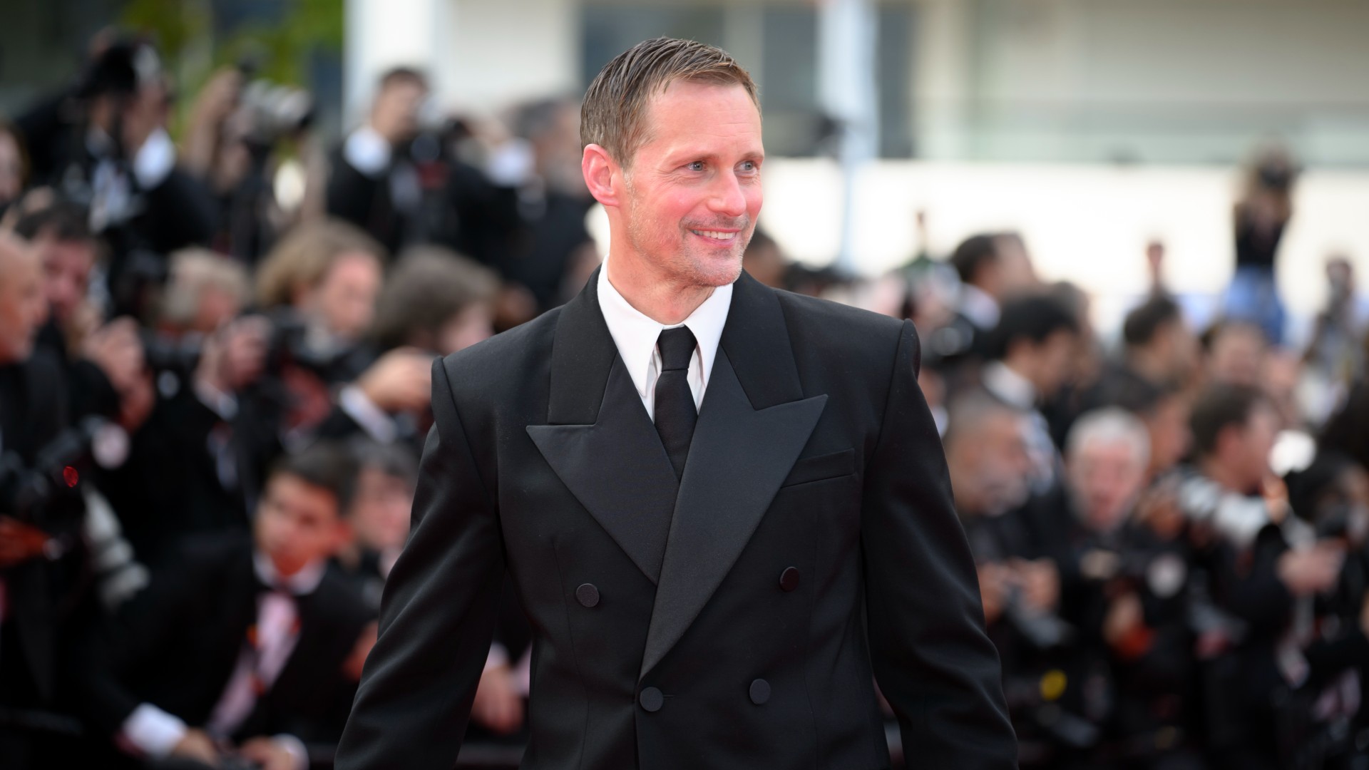 a white man wearing a black tuxedo posing in front of paparazzi on a red carpet 