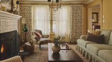 a warm yellow living room with floral skirted couch, floral curtains, vintage wooden furniture and a lit tiled fire
