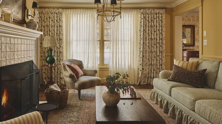 a warm yellow living room with floral skirted couch, floral curtains, vintage wooden furniture and a lit tiled fire
