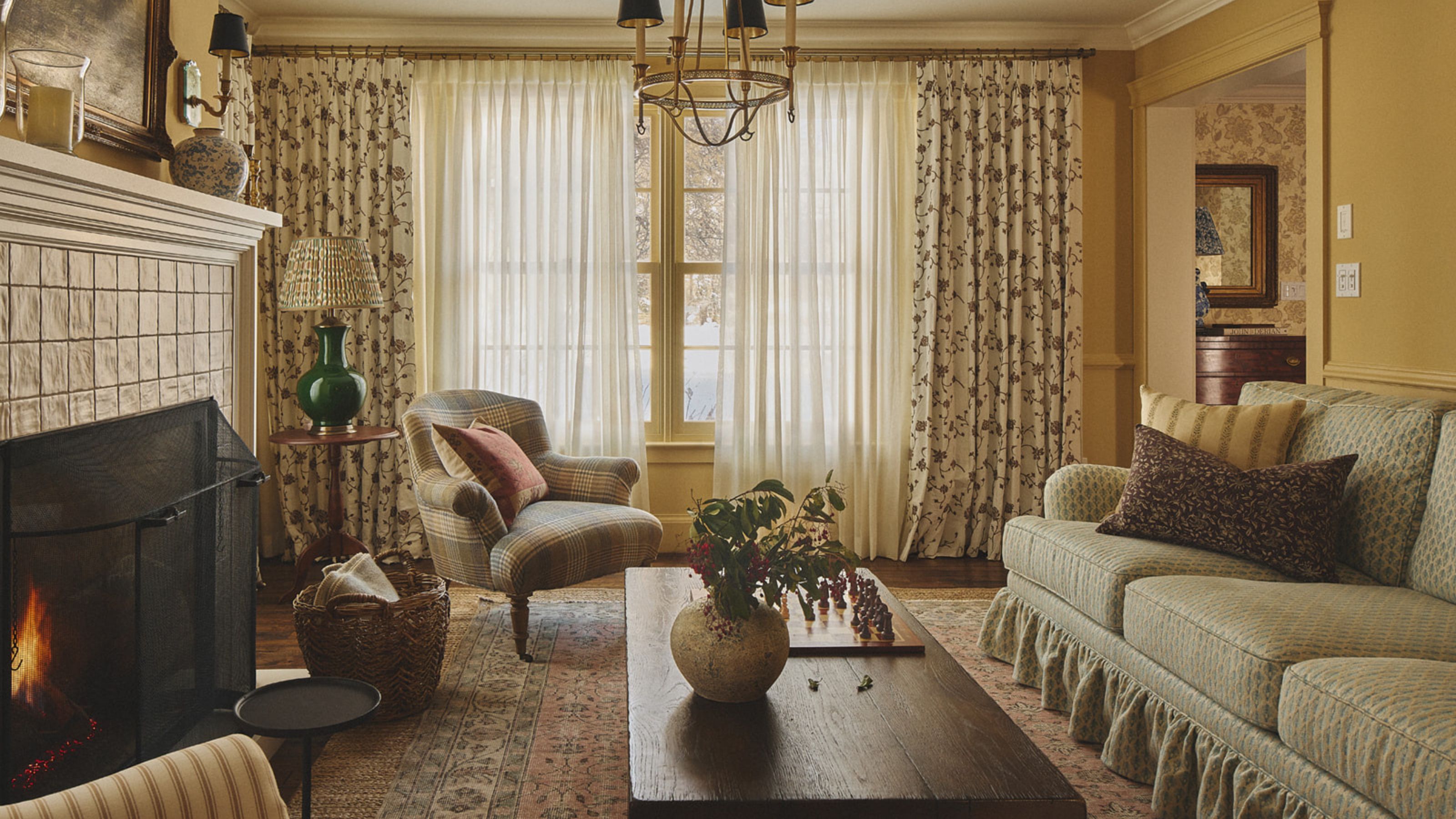a warm yellow living room with floral skirted couch, floral curtains, vintage wooden furniture and a lit tiled fire