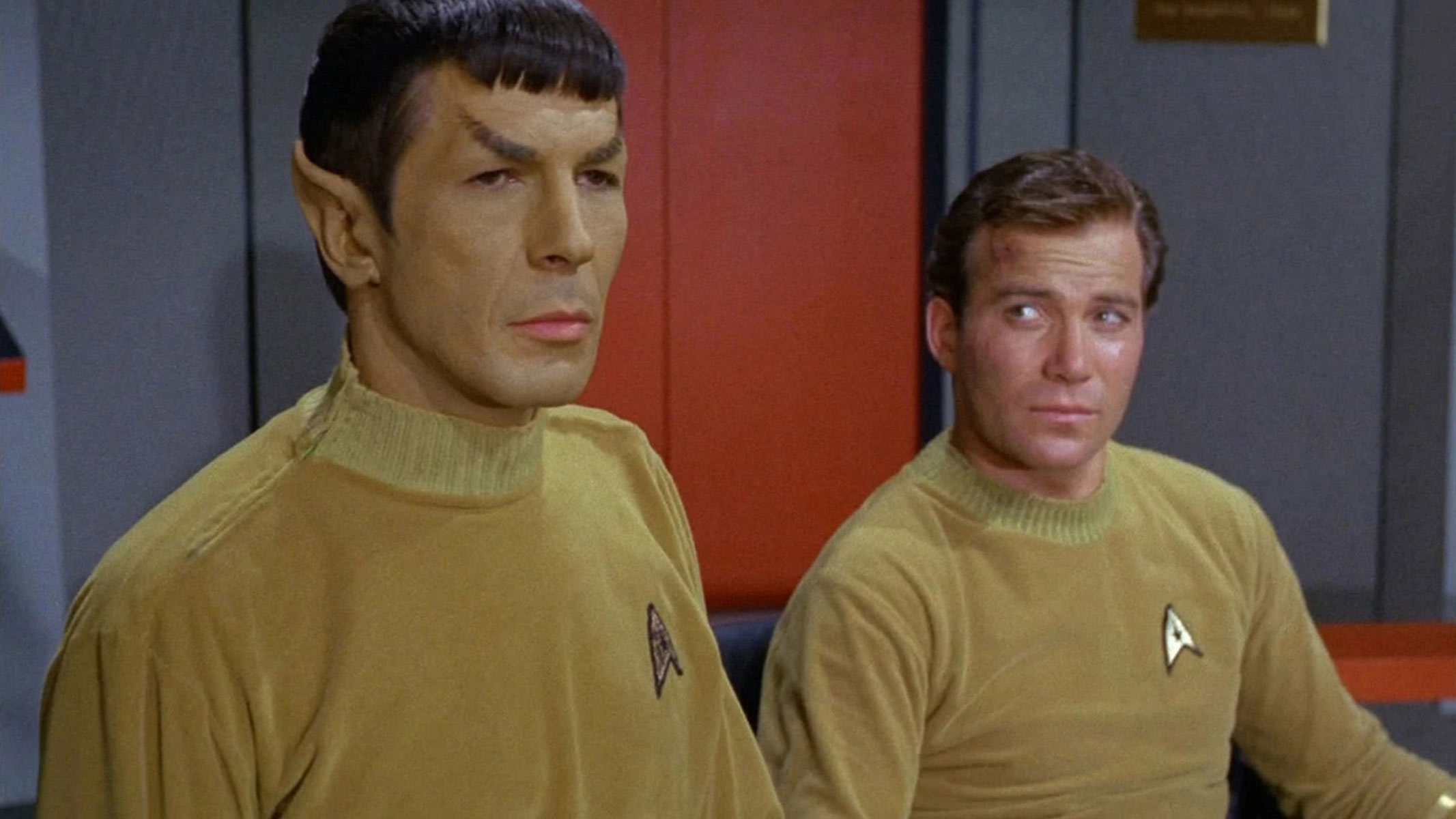 Two men, one with pointy ears are sat with mustard-colored jumpers on and an emblem on the lapel. 